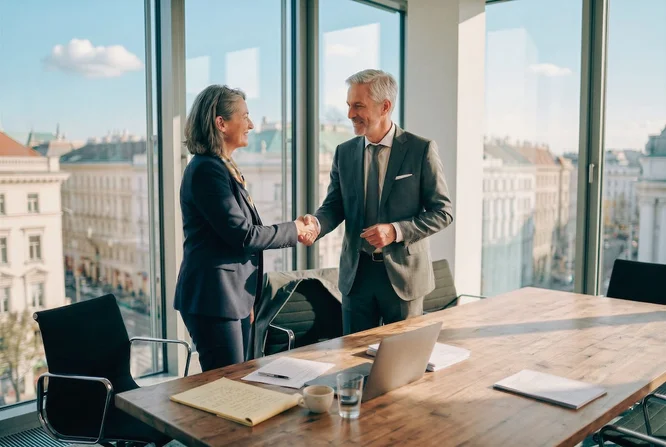 Zwei Erwachsene in Businesskleidung schütteln sich in einem hellen, modernen Büro mit großen Fenstern an einem hölzernen Konferenztisch die Hände. Der Blick auf die Stadtlandschaft vermittelt ein professionelles oder geschäftliches Ambiente. Auf dem Tisch liegen Dokumente, ein Laptop und Kaffeetassen, was auf ein formelles Treffen oder eine Vereinbarung hindeutet. Die Atmosphäre ist professionell und kooperativ, und natürliches Tageslicht erhellt den Raum.