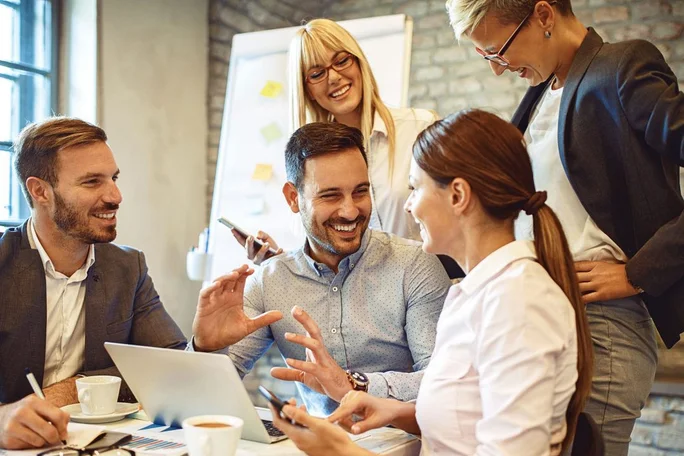 Eine Gruppe von Fachleuten führt in einem modernen Büro ein angeregtes Gespräch. Laptops, Smartphones, Kaffeetassen und ein Whiteboard mit Diagrammen im Hintergrund prägen die Arbeitsumgebung. Die Atmosphäre vermittelt Teamgeist und Produktivität in einem ungezwungenen, aber dennoch professionellen Arbeitsumfeld.