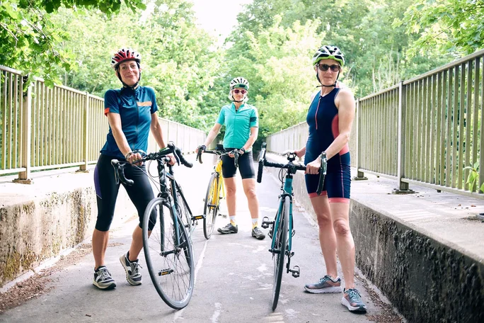Drei Radfahrer in Sportkleidung stehen mit ihren Rennrädern auf einer sonnenbeschienenen Brücke, umgeben von üppigem Grün. Die Szenerie lässt auf eine Freizeitaktivität im Freien an einem klaren Tag schließen. Die Gruppe trägt Helme und Radbekleidung, was Sicherheit und Fitness unterstreicht.