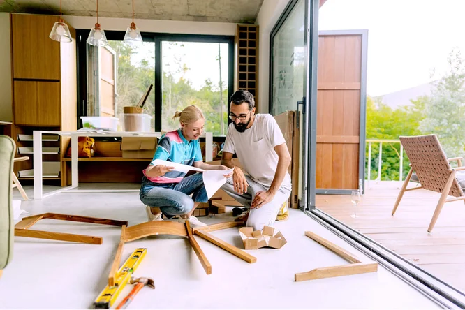 Ein Mann und eine Frau montieren in einem hellen, modernen Haus mit großen Glastüren zur Terrasse Holzmöbel. Das Ambiente ist lichtdurchflutet, mit Holzelementen gestaltet und entspannt. Werkzeug und Holzteile, darunter eine gelbe Wasserwaage, liegen verstreut auf dem Boden. Im Hintergrund sieht man Grünpflanzen und Sitzgelegenheiten im Freien.