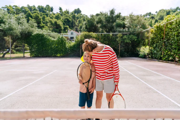 Eine Frau in rot-weiß gestreiftem Hemd und Shorts steht mit einem Kind auf einem Tennisplatz im Freien. Beide halten Tennisschläger, und der Platz ist von Grün und Bäumen umgeben. Die Umgebung ist hell und sonnig und schafft eine fröhliche und entspannte Atmosphäre.