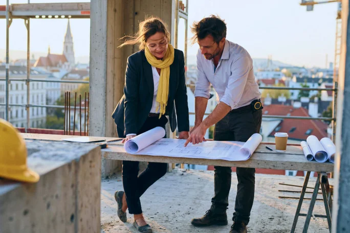 Zwei Erwachsene, ein Mann und eine Frau, studieren Baupläne auf einem Tisch an einer Baustelle. Die Szenerie ist urban geprägt; im Hintergrund sind Stadtgebäude und ein Kirchturm zu sehen. Das natürliche Tageslicht und die industriellen Elemente vermitteln eine professionelle, kooperative Atmosphäre. Ein gelber Bauhelm und eine Kaffeetasse auf dem Tisch unterstreichen das Arbeitsumfeld.