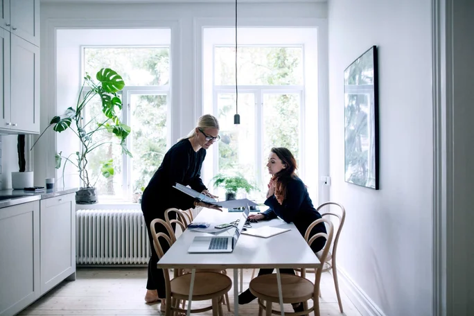 Zwei Frauen arbeiten gemeinsam an einem weißen Esstisch in einer hellen, modernen Küche. Große Fenster lassen viel Tageslicht herein, und die minimalistische Einrichtung mit Grünpflanzen sorgt für einen einladenden Raum. Ein Laptop und Dokumente liegen auf dem Tisch und deuten auf eine berufliche oder kreative Zusammenarbeit hin.
