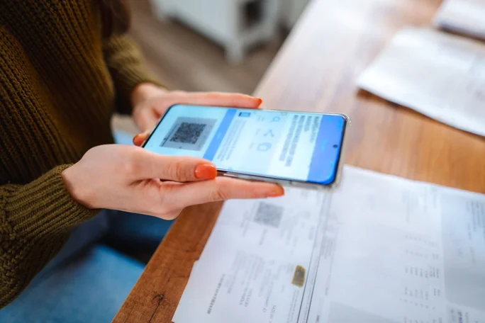 Nahaufnahme einer Person, die mit einem Smartphone einen QR-Code auf dem Bildschirm scannt. Die Szene spielt sich offenbar in einem Innenraum ab; auf einem Holztisch liegen Dokumente und Papiere. Die Person trägt einen grünen Pullover und hat orange lackierte Fingernägel, was einen Farbtupfer setzt.