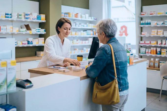 Ein Apotheker im weißen Kittel bedient eine ältere Dame am Apothekenschalter. Die Szene spielt in einer modernen Apotheke mit Regalen voller Medikamente und anderer Produkte. Die Interaktion wirkt professionell und freundlich; die ältere Dame trägt eine hellbraune Handtasche. Die Atmosphäre ist insgesamt ruhig und beruhigend, die Szene ist in neutralen und sanften Farbtönen gehalten.
