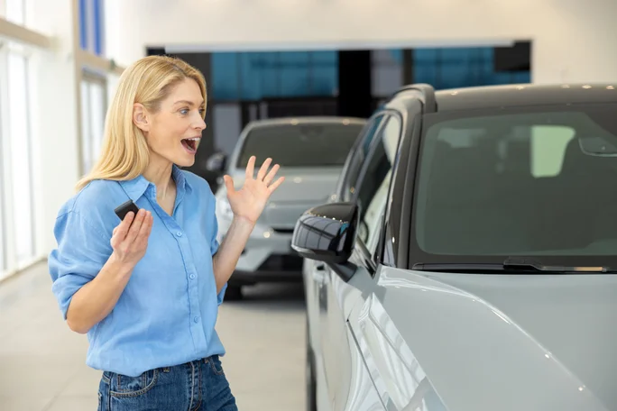 Eine glückliche, hübsche, langhaarige Blondine steht neben einem neuen Auto und wirkt vergnügt und aufgeregt.