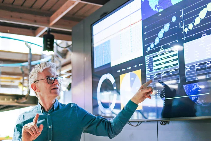 Eine Person deutet auf einen großen Bildschirm, auf dem verschiedene Datenvisualisierungen wie Diagramme, Grafiken und numerische Kennzahlen angezeigt werden. Die Umgebung wirkt wie ein modernes Büro oder ein Technologie-Arbeitsplatz mit Elementen im Industriestil. Die Visualisierungen umfassen Kreisdiagramme, Balkendiagramme und globale Datendarstellungen und betonen so den Fokus auf Analytik und Technologie.
