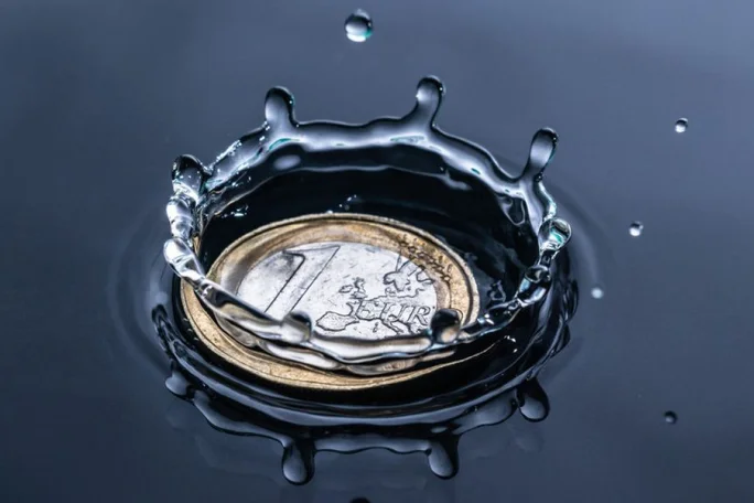 A single Euro coin is captured mid-splash as it lands in water, creating a dramatic ripple effect. The setting emphasizes fluid motion and reflective surfaces, with a close-up view highlighting the coin's intricate design. The image conveys a sense of precision and dynamic energy.