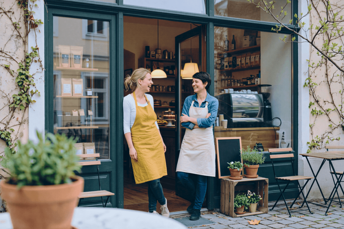Zwei Erwachsene in Schürzen unterhalten sich am Eingang eines kleinen Cafés. Der Außenbereich mit Topfpflanzen lädt zum Verweilen ein und wirkt einladend. Durch große Fenster kann man ins Innere sehen, wo Regale und eine Kaffeemaschine stehen. Die Atmosphäre ist entspannt und freundlich, mit viel Tageslicht und warmen Erdtönen. Auf dem Bild sind keine Texte oder Zahlen zu erkennen.