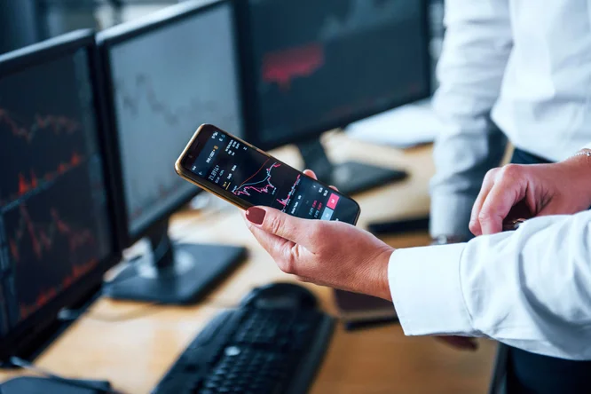 Nahaufnahme einer Person mit einem Smartphone, auf dem Börsencharts und -daten angezeigt werden. Die Szene spielt in einem Büro mit mehreren Monitoren, auf denen im Hintergrund Finanzgrafiken zu sehen sind. Die Person trägt formelle Kleidung, was auf einen professionellen Kontext hindeutet. Auf dem Smartphone-Bildschirm sind deutlich rote und grüne Chartgrafiken zu sehen.