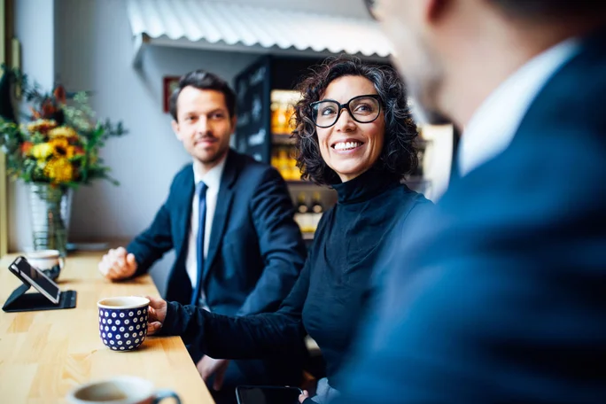 Eine Gruppe von Geschäftsleuten sitzt an einem Holztisch in einem Café und unterhält sich. Auf dem Tisch stehen Kaffeetassen und eine Smartphone-Halterung, im Hintergrund ist ein Blumenarrangement zu sehen. Die Atmosphäre ist ungezwungen und dennoch professionell, das natürliche Licht unterstreicht die warmen Farbtöne des Raumes.