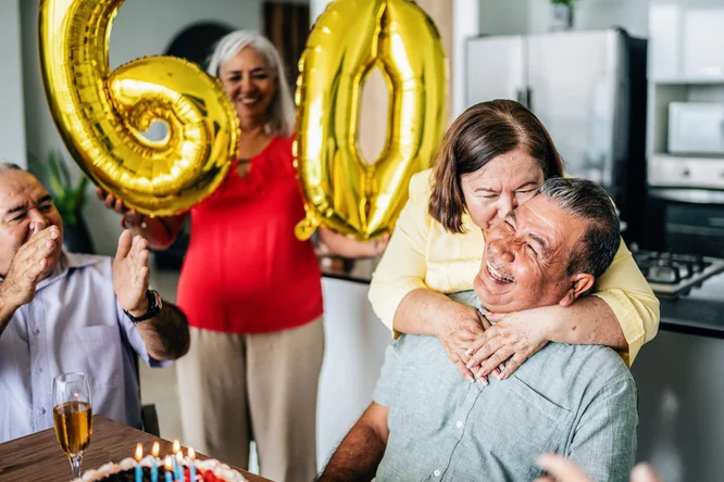 Eine fröhliche Feier zum 60. Geburtstag in privater Atmosphäre. Goldene „60“-Luftballons schmücken den Raum und verleihen ihm eine festliche Note. Auf dem Tisch stehen eine Torte mit brennenden Kerzen und ein Glas Champagner, was die Feierlaune unterstreicht. Im Hintergrund ist eine moderne Küche zu sehen.