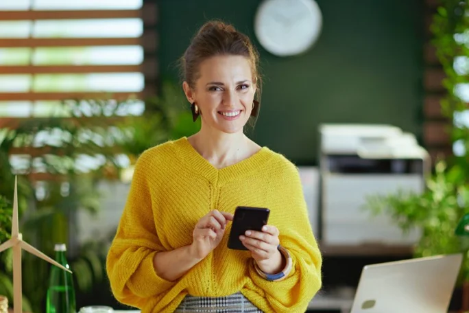 Eine Frau in einem leuchtend gelben Pullover hält in einem modernen Büro ein Smartphone in der Hand. Im Hintergrund sind Grünpflanzen, eine Uhr und Bürogeräte zu sehen, die eine frische und professionelle Atmosphäre schaffen. Auf dem Schreibtisch stehen ein kleines Windturbinenmodell und eine Wasserflasche, die das umweltfreundliche Thema unterstreichen.
