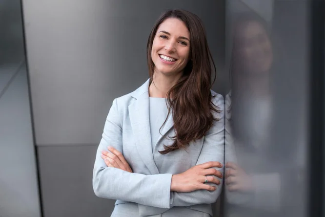 Eine Frau steht selbstbewusst mit verschränkten Armen da, in einem hellgrauen Blazer und dazu passender Kleidung. Sie posiert vor einer modernen Metallwand in einer urbanen Umgebung im Freien. Das Bild vermittelt Professionalität und Souveränität.