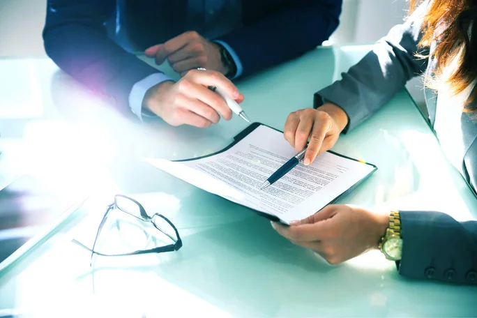 Zwei Geschäftsleute sitzen an einem Glastisch, besprechen ein Dokument und unterzeichnen es. Die Szenerie erinnert an ein helles Bürogebäude. Auf dem Tisch liegen ein Stift, eine Brille und ein formeller Vertrag.
