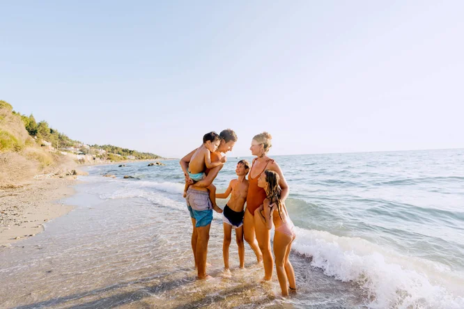 Eine fünfköpfige Familie, bestehend aus Erwachsenen und Kindern, verbringt gemeinsam Zeit an einem malerischen Strand. Strahlend blauer Himmel, sanfte Wellen und ein feiner Sandstrand prägen die Atmosphäre. Die Stimmung ist fröhlich und entspannt, warme Farbtöne dominieren. Die Gruppe trägt Badekleidung, was auf einen unbeschwerten und fröhlichen Ausflug ans Meer hindeutet.