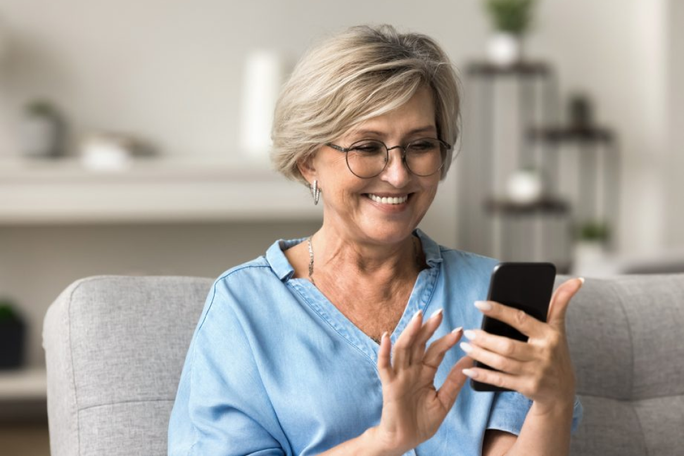 An older woman is seated on a gray couch in a modern living room, holding and interacting with a smartphone. She is wearing a light blue denim-style shirt, and the background features minimalistic decor with shelves and plants. The setting suggests a casual and relaxed atmosphere.