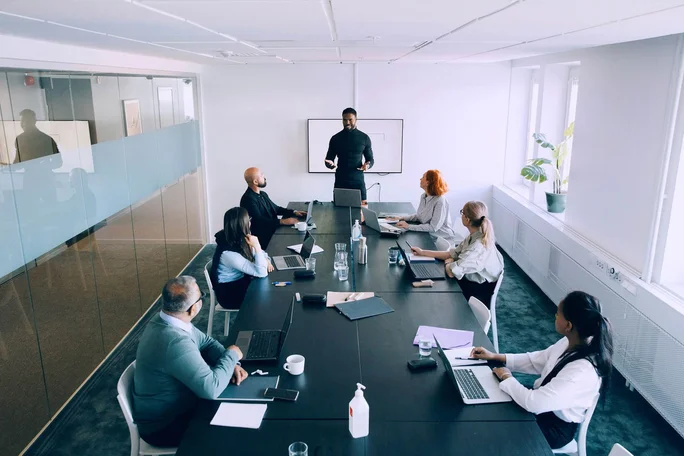 Ein professionelles Umfeld mit einer vielfältigen Gruppe von Personen, die um einen Konferenztisch sitzen und ein Geschäftstreffen abhalten. Der/Die Präsentator/in steht neben einem Whiteboard und spricht zum Team. Der Raum ist modern gestaltet, mit großen Fenstern, viel Tageslicht und minimalistischer Einrichtung.