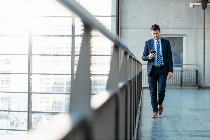Ein Mann in formeller Kleidung – marineblauer Blazer, gestreifte Krawatte und Jeans – geht durch einen hellen, modernen Flur. Er hält ein Smartphone in der Hand, was auf die Nutzung von Technologie oder Kommunikation hindeutet. Große Glasfenster und die Architektur im Industriestil schaffen eine professionelle und zeitgemäße Atmosphäre.