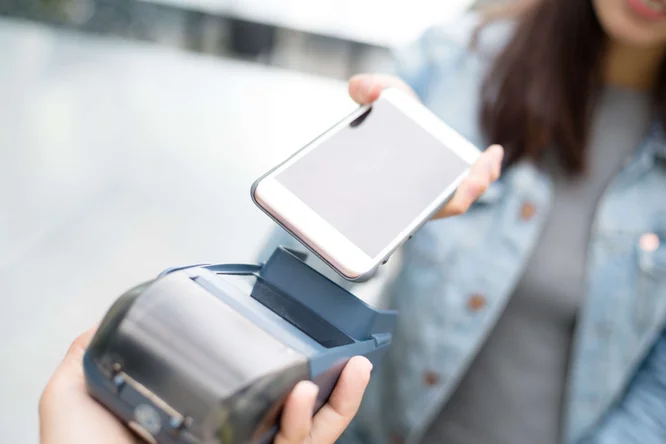Eine Person hält ein Smartphone an ein drahtloses Kartenlesegerät und demonstriert eine kontaktlose Zahlungstransaktion. Die Szene spielt sich im Freien ab; die Person trägt eine lässige Jeansjacke. Der Fokus des Bildes liegt auf den Händen und Geräten und unterstreicht so die moderne Technologie und den Komfort. Auf den Geräten sind keine Texte oder Zahlen sichtbar.