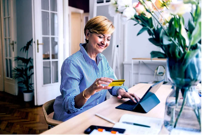 Eine Frau sitzt an einem Holzschreibtisch und hält eine gelbe Kreditkarte in der Hand, während sie ein Tablet benutzt. Es ist ein helles, modernes Homeoffice mit Blumen und Schreibwaren auf dem Schreibtisch. Die Atmosphäre ist professionell und ordentlich, und natürliches Licht strömt durch die Fenster.