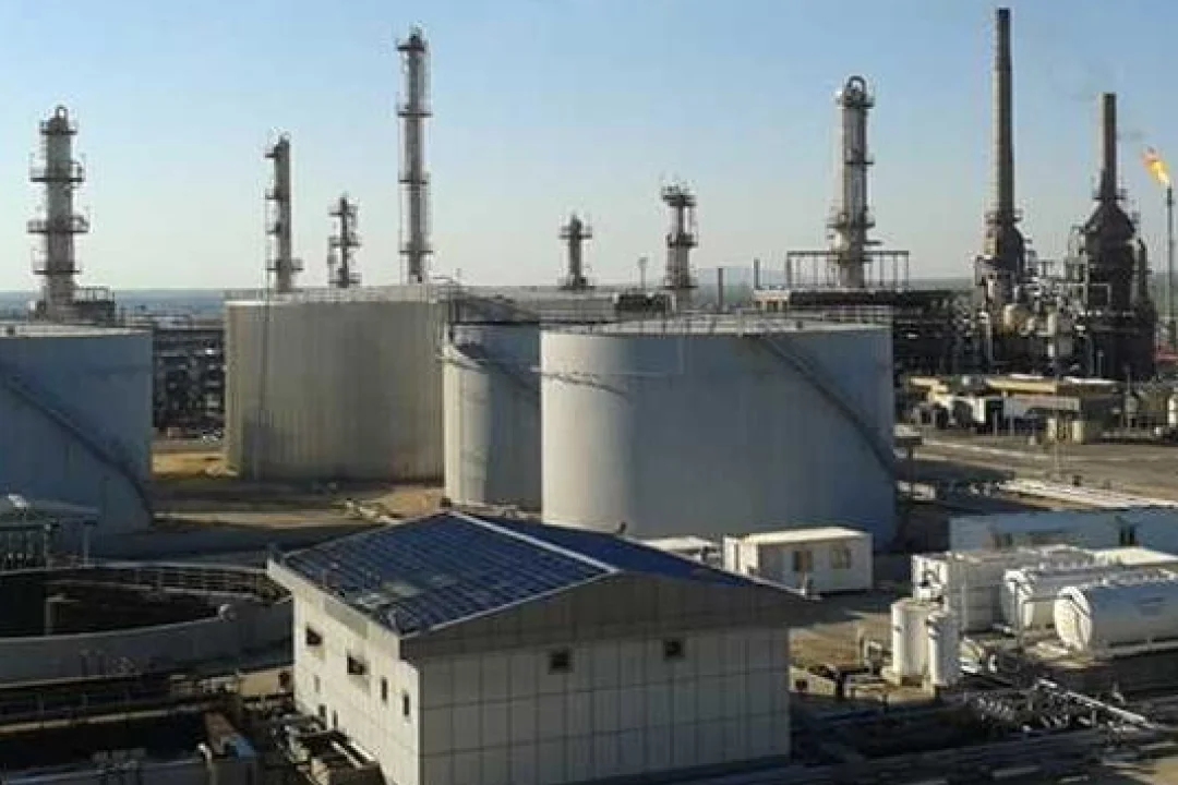 Eine große industrielle Ölraffinerie mit mehreren zylindrischen Lagertanks und hohen Schornsteinen. Die Szene spielt im Freien unter klarem, blauem Himmel; die Industrieanlagen und -geräte sind gut zu erkennen. Die Anlage scheint in Betrieb zu sein; an einem der Schornsteine ​​ist eine Flamme sichtbar.
