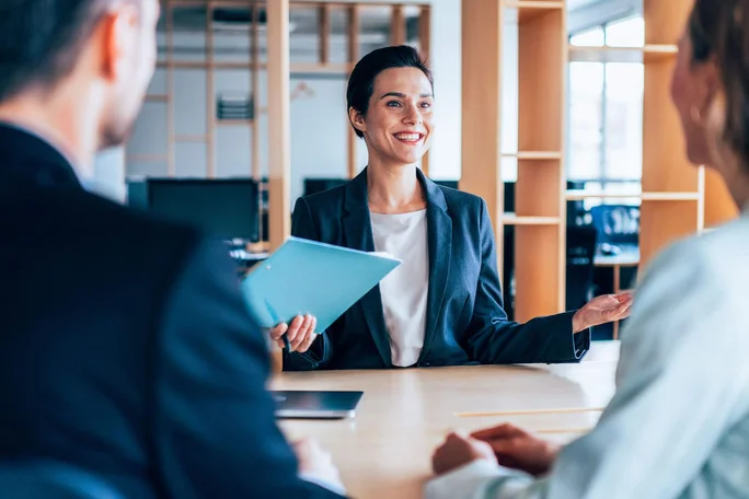Eine Frau in Businesskleidung sitzt an einem Schreibtisch und unterhält sich mit zwei Personen ihr gegenüber in einem modernen Büro. Sie hält eine blaue Mappe und gestikuliert selbstbewusst, was eine positive und kooperative Atmosphäre signalisiert. Im Hintergrund sind Holzregale und Computermonitore zu sehen, die das professionelle Ambiente unterstreichen. Die Stimmung ist insgesamt professionell und einladend.