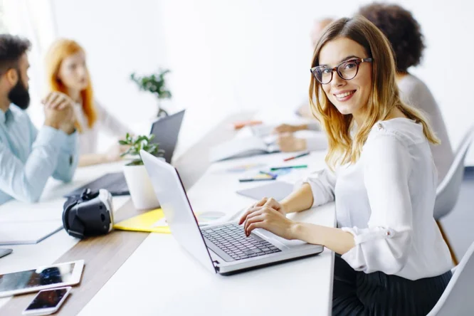 Eine Gruppe von Fachleuten arbeitet in einem hellen, modernen Büro zusammen. Im Fokus steht eine Frau, die auf einem Laptop tippt, umgeben von Kollegen, die sich angeregt unterhalten. Der Arbeitsplatz ist minimalistisch eingerichtet; Pflanzen und Büromaterialien stehen auf dem Tisch. Die Atmosphäre wirkt kooperativ und produktiv.