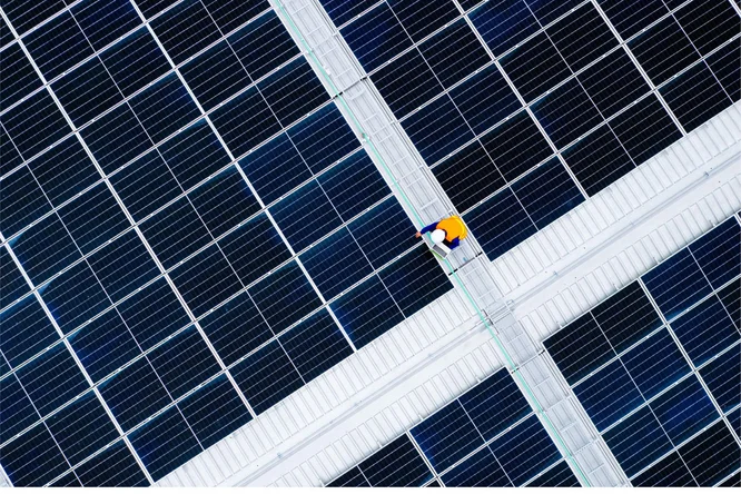 Eine Person in gelbem Hemd und blauer Hose geht auf einem mit Solarmodulen bedeckten Dach. Es handelt sich offenbar um ein Industrie- oder Gewerbegebäude mit Schwerpunkt auf erneuerbarer Energie. Das Bild unterstreicht die klare, geometrische Anordnung der Module und das Thema nachhaltige Energie.