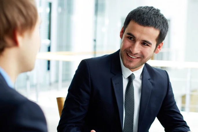 Zwei Personen in formeller Geschäftskleidung führen in einem modernen Büro ein professionelles Gespräch. Im Hintergrund dominieren helles Tageslicht und zeitgenössische Architektur. Im Mittelpunkt stehen Zusammenarbeit und Kommunikation im Unternehmensumfeld.