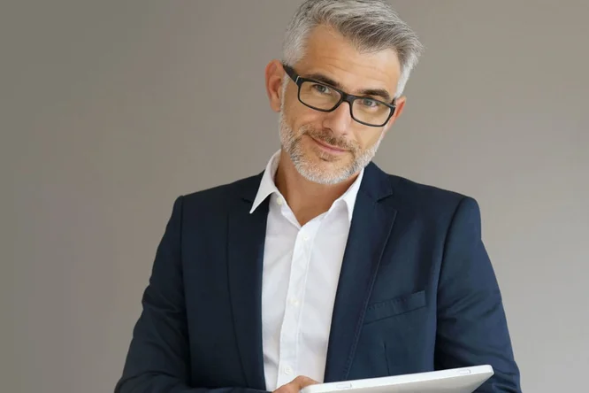 Ein Mann in einem dunkelblauen Anzug und weißem Hemd hält in einem neutralen Innenraum ein Tablet in der Hand. Der schlichte Hintergrund unterstreicht die professionelle Kleidung und die moderne Technologie. Das Bild vermittelt mit seiner klaren und einfachen Gestaltung eine geschäftliche Atmosphäre.