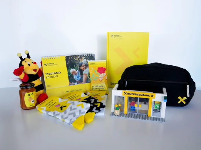 Geschenke für den Weltspartag vor einer weißen Wand. Ein Kalender, Socken, ein Notizbuch und ein Sumsi Kuscheltier
