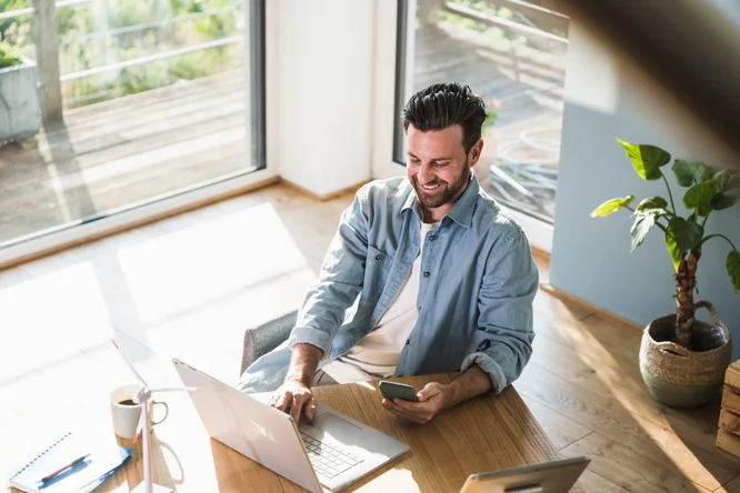 Mann in sonnendurchflutetem Raum vor Laptop mit Smartphone in der Hand