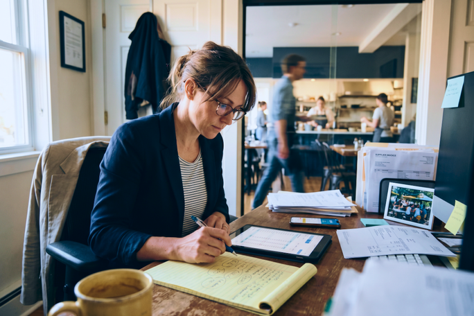 Eine Frau sitzt an einem Schreibtisch und schreibt auf einem gelben Notizblock in einem hellen, modernen Büro. Auf dem Schreibtisch befinden sich Unterlagen, eine Kaffeetasse und ein Tablet. Im verschwommenen Hintergrund sind vorbeigehende Personen und ein Küchenbereich zu sehen. Die Szene vermittelt eine geschäftige, produktive Atmosphäre mit natürlichem Tageslicht und ordentlich arrangierten Materialien. Weder Text noch Zahlen sind auf dem Bild eindeutig erkennbar.