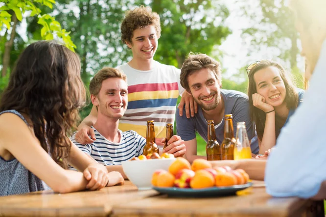 Eine Gruppe von Menschen genießt ein gemütliches Beisammensein im Freien an einem Holztisch. Üppiges Grün und Sonnenlicht schaffen eine warme und entspannte Atmosphäre. Getränkeflaschen und eine Schale mit frischen Aprikosen stehen gut sichtbar auf dem Tisch und unterstreichen die ungezwungene Stimmung.