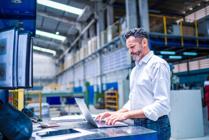 Ein Mann arbeitet in einer Fabrikhalle an einem Laptop. Sein Arbeitsplatz ist mit mehreren Bildschirmen, einem Tablet und verschiedenen Geräten ausgestattet. Hohe Decken, Metallkonstruktionen und ordentlich organisierte Arbeitsplätze deuten auf eine Produktionsstätte hin.