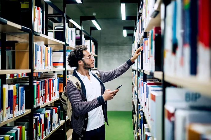 Ein junger Mann mit Rucksack stöbert in einer hellen, modernen Bibliothek. In der einen Hand hält er sein Smartphone, mit der anderen greift er nach einem Buch im Regal. Die Bibliothek besticht durch ordentliche Regale voller bunter Bücher und einen grünen Teppichboden, die eine konzentrierte und zeitgemäße Atmosphäre schaffen.