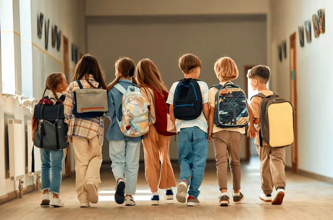 7 Kinder mit Rucksäcken gehen gemeinsam Gang eines Schulgebäudes entlang