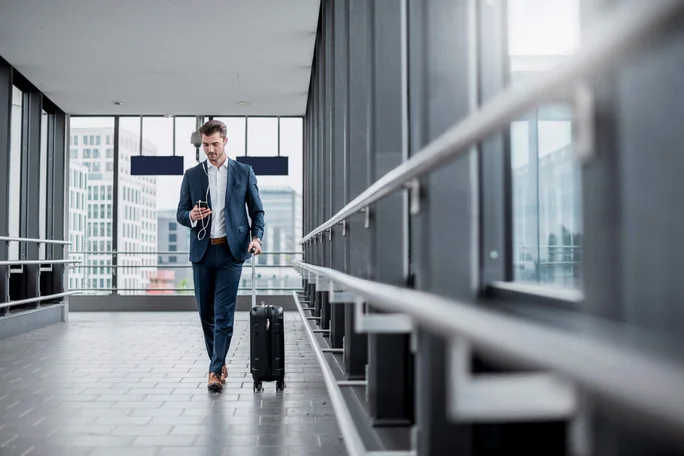 Ein Mann im blauen Anzug geht mit Smartphone und Kopfhörern durch einen modernen Flughafenkorridor. Er hat einen Rollkoffer dabei, was auf eine Reise oder einen Arbeitsweg hindeutet. Große Fenster geben den Blick auf die Stadtarchitektur im Hintergrund frei.