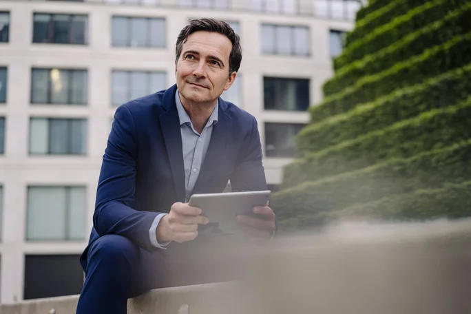Ein Mann in einem dunkelblauen Anzug sitzt im Freien und hält ein Tablet in der Hand. Die Umgebung zeichnet sich durch moderne Architektur mit großen Fenstern und einer terrassenförmig angelegten Grünanlage im Hintergrund aus. Die Szene vermittelt eine professionelle und urbane Atmosphäre.
