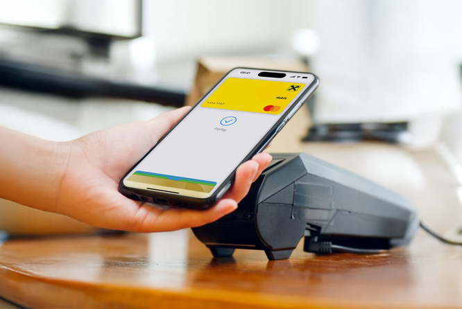 Eine Hand hält ein Smartphone mit der Benutzeroberfläche einer Zahlungs-App neben einem Kartenlesegerät für kontaktloses Bezahlen. Der Bildschirm zeigt eine gelb-weiße Oberfläche mit einem gut sichtbaren Logo und dem Wort „Bezahlen“. Die Szene spielt sich offenbar in einem Innenraum auf einer Holzoberfläche ab und unterstreicht so moderne Technologie und Komfort.
