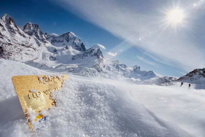Eine goldene Kreditkarte, die teilweise im Schnee vergraben ist, steht im Mittelpunkt des Bildes. Die Szenerie zeigt eine atemberaubende, schneebedeckte Bergkette unter strahlend blauem Himmel. Im Hintergrund sind drei kleine Gestalten zu sehen, die durch den Schnee laufen und dem Bild Tiefe und Dynamik verleihen.