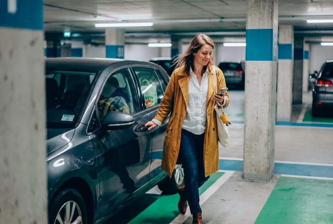 Frau mit Smartphone in der Hand verlässt ihr Auto in einer Parkgarage 