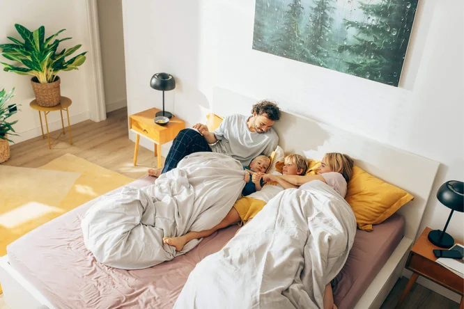 Eine vierköpfige Familie – zwei Erwachsene und zwei Kinder – entspannt sich gemeinsam auf einem Bett in einem hellen, modernen Schlafzimmer. Warme Farbtöne, natürliches Licht und Zimmerpflanzen schaffen eine gemütliche und einladende Atmosphäre. Ein Wandbild mit Waldmotiven verleiht dem Raum einen naturnahen Touch.