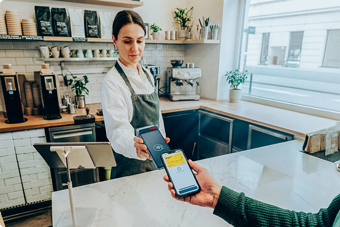 In einem modernen Café hält ein Barista ein Zahlungsterminal in der Hand, während ein Kunde mit seinem Smartphone bezahlt. Das Café besticht durch ein helles, sauberes Interieur mit weißen Fliesen und Holzelementen. Auf dem Smartphone-Bildschirm ist eine Zahlungs-App mit einer gut sichtbaren gelben Benutzeroberfläche zu sehen. Die Szene vermittelt ein modernes, effizientes und bargeldloses Zahlungserlebnis.