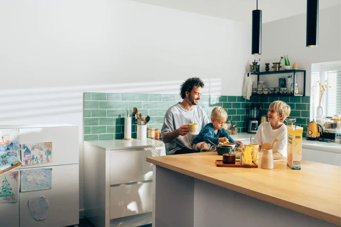 Eine Familie genießt gemeinsam ihr Frühstück in einer hellen, modernen Küche mit grünen Fliesen als Spritzschutz und Arbeitsplatten aus Holz. Verschiedene Frühstückszutaten, ein Saftkarton und eine gemütliche Atmosphäre prägen das Bild. Kinderzeichnungen am Kühlschrank verleihen dem Raum eine persönliche Note. Die Küche ist dank Tageslicht und Pendelleuchten gut beleuchtet.