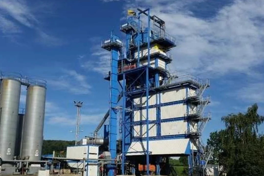 Eine große Industrieanlage mit markanter blauer Stahlkonstruktion und mehreren Ebenen. Die Anlage befindet sich im Freien unter strahlend blauem Himmel, in der Nähe sind Lagersilos zu sehen. Umgebendes Grün und Bäume bilden einen Kontrast zum industriellen Erscheinungsbild.