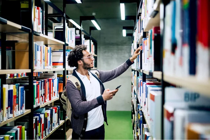 Ein junger Mann mit Rucksack stöbert in einer hellen, modernen Bibliothek. In der einen Hand hält er sein Smartphone, während er nach einem Buch im Regal greift. Die Bibliothek besticht durch ordentliche Regale voller bunter Bücher und einen grünen Teppichboden. Die Atmosphäre ist ruhig und konzentriert – ideal zum Lernen oder Recherchieren.