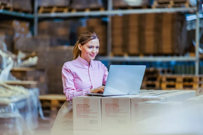 Eine Person in einem rosa Hemd arbeitet an einem Laptop, der auf gestapelten Kisten in einer Lagerhalle steht. Im Hintergrund sind Regale mit Paletten und verpackten Waren zu sehen. Die Szene vermittelt ein industrielles und organisiertes Umfeld, ideal für Logistik- oder Bestandsverwaltungstätigkeiten.
