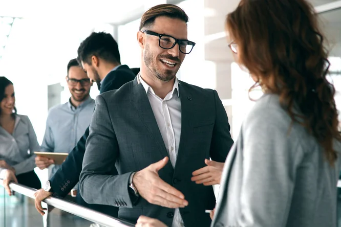 Eine Gruppe von Geschäftsleuten führt in einem modernen Büro eine formelle Diskussion. Das Bild zeigt Personen in Businesskleidung, darunter Anzüge und Brillen, die sich in der Nähe eines Geländers unterhalten. Die helle und moderne Atmosphäre deutet auf eine Firmenveranstaltung oder ein Networking-Event hin.